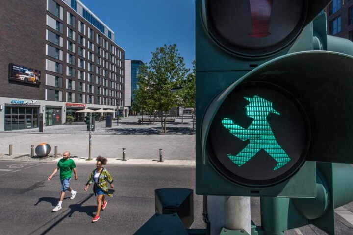 In the foreground, a pedestrian traffic light is switched to green. In the background, a man and a woman are walking across the street