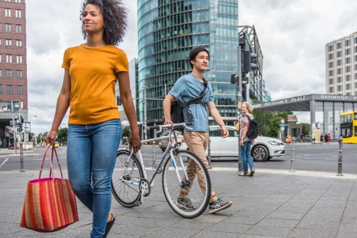 Eine junge Frau läuft im Vordergrund, dahinter läuft ein Mann mit Fahrrad, im Hintergrund ein Auto vor dem DB Tower am Potsdamer Platz