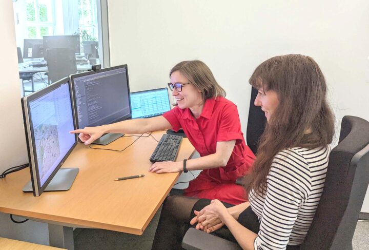 Zwei Frauen sitzen an Schreibtisch und schauen auf Computerbildschirme mit Analysen.