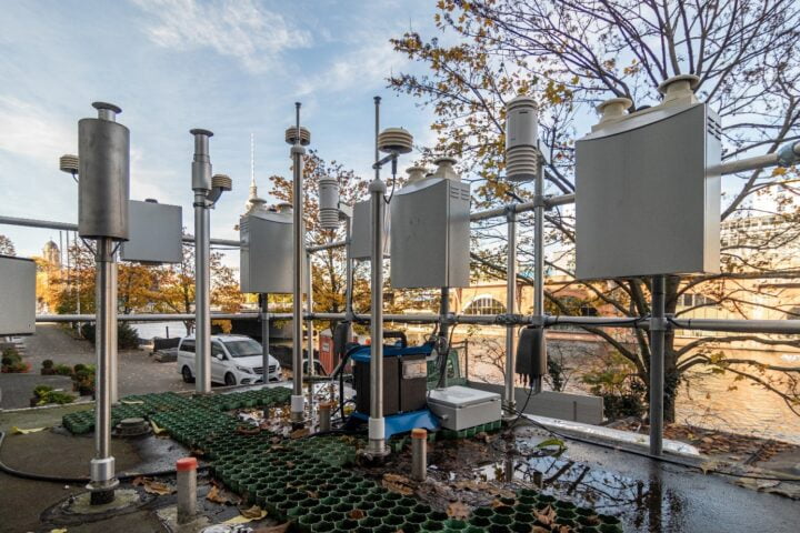 Das Foto zeigt Luftqualitätssensorsysteme in Form von mehreren geschlossenen grauen Metallkästen, die Briefkästen ähneln. Zusammen mit mehreren Rohren und pilzartigen Lamellensensoren sind sie an einem Gerüst oberhalb des Bodens angebracht. Im Hintergrund ist der Berliner Fernsehturm zu erkennen.