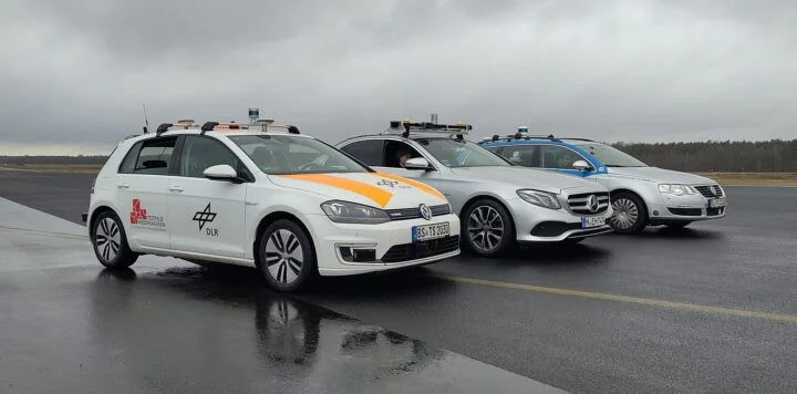 Auf dem Foto sind drei Autos mit kleinen Aufbauten in Zylinderform auf den jeweiligen Dächern zu sehen. Sie stehen nebeneinander in gleicher Ausrichtung unter bewölktem Himmel auf erkennbar feuchtem Asphalt. AUf dem vordersten Auto ist seitlich auf der Beifahrertür das Logo des DLR zu erkennen. Hinten im Bild ist am Ende der freien Fläche Wald zu sehen.